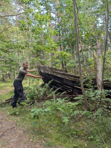 Nico zeigt auf ein altes Boot, welches in einem Wald steht und sehr verrottet ist